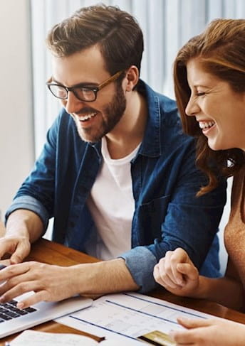 A happy couple smiling and laughing while using a laptop together at a table