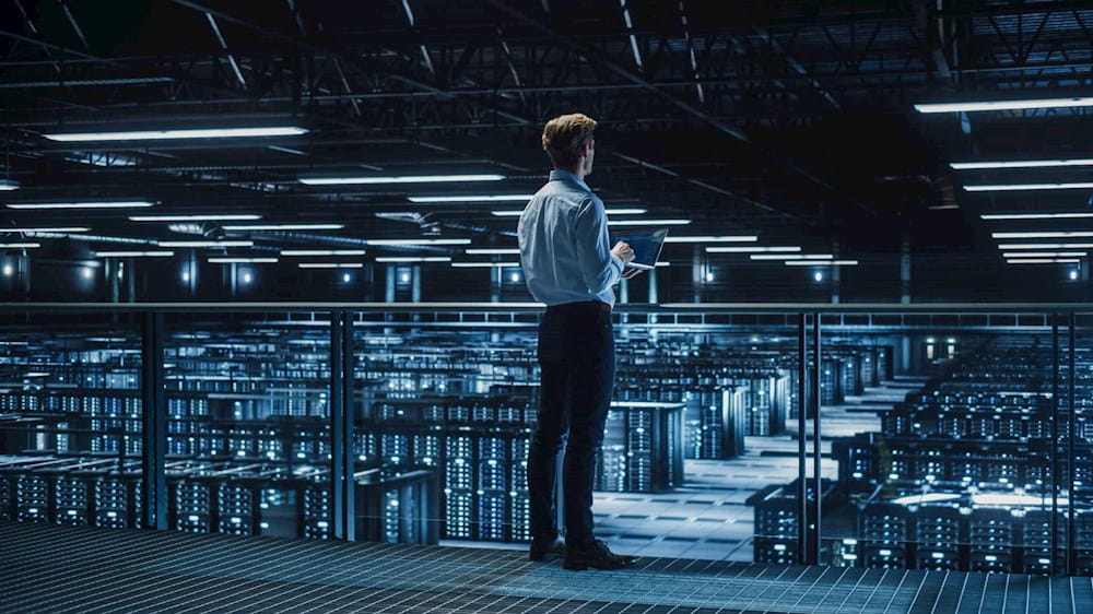 Uomo in piedi con un pc in mano si trova dentro una grande stanza piena di computer