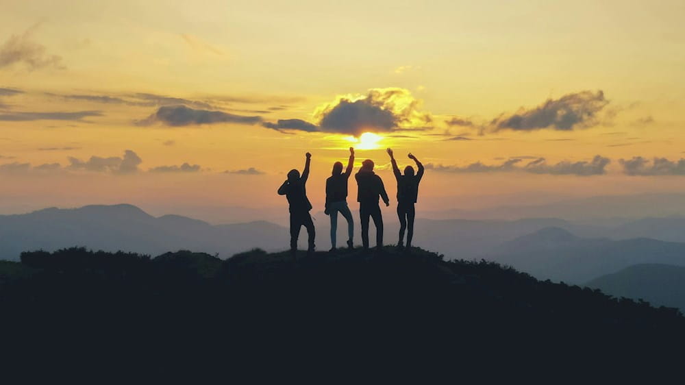 sagome in lontananza di 4 persone che festeggiano l'essere arrivati in cima a una collina al tramonto