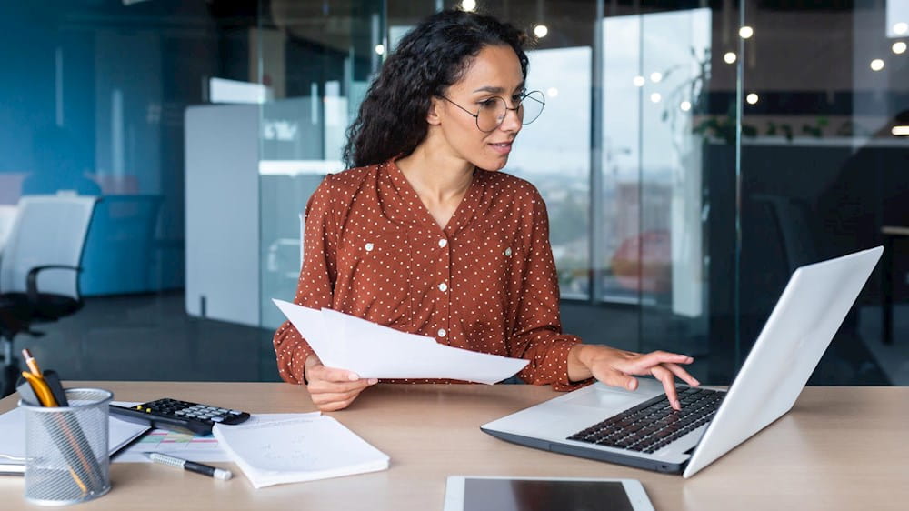 Una donna in ufficio seduta alla scrivania lavora al computer e tiene un foglio in mano