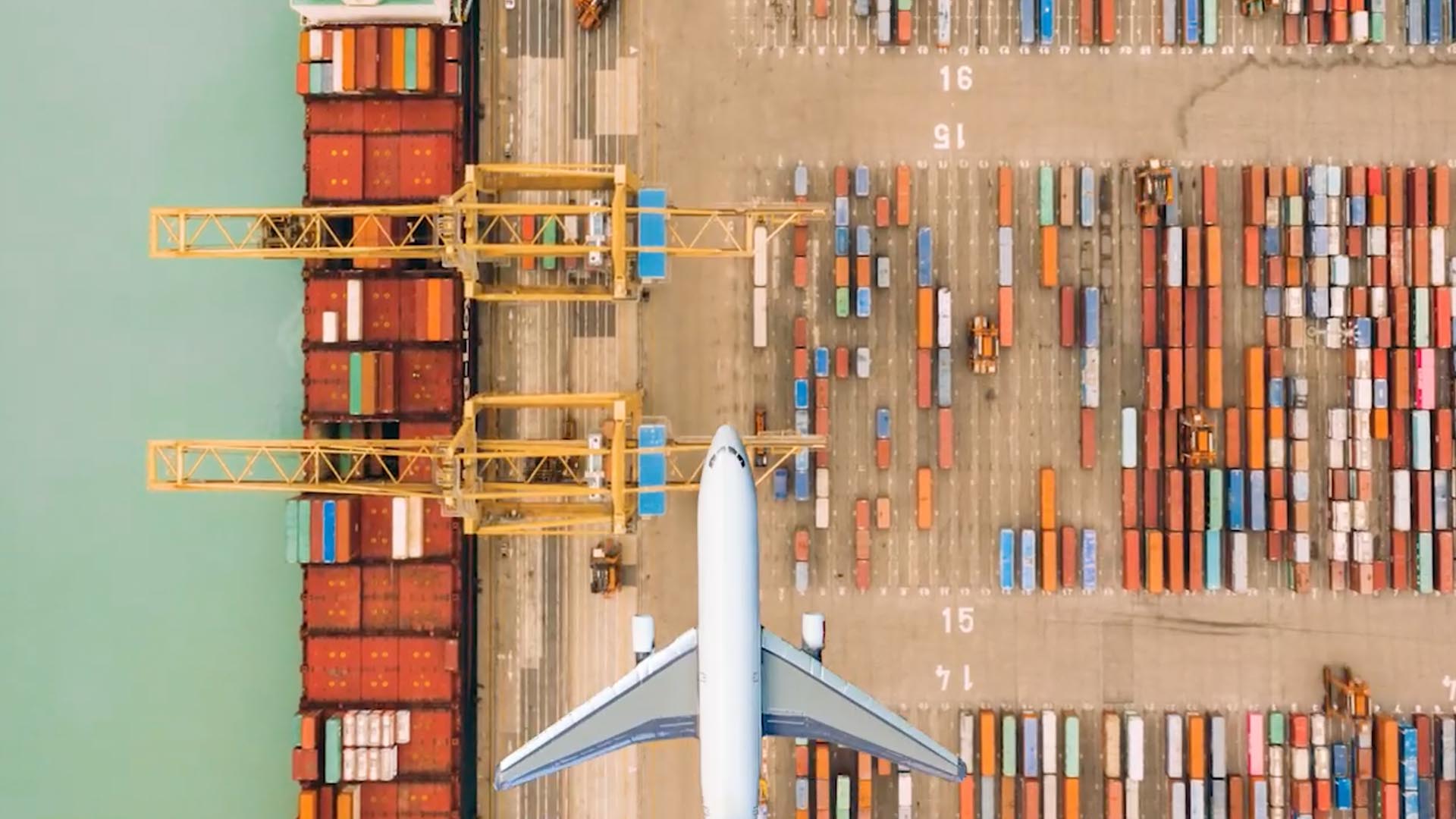 A plane flying over shipping containers and a ship on a dockyard
